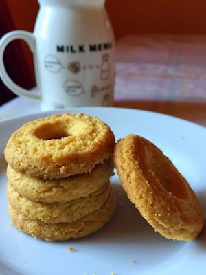 A white plate holds a stack of round, golden-brown cookies with a crumbly texture. In the background, there is a ceramic mug featuring milk-themed imagery and text.