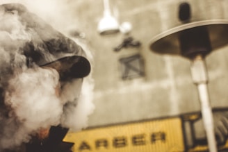 A person wearing a cap is partially obscured by thick smoke, standing in front of an industrial background with pipes and vents. The scene is somewhat blurred, giving it a mysterious and atmospheric feel.