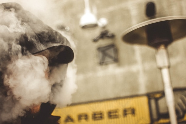 A person wearing a cap is partially obscured by thick smoke, standing in front of an industrial background with pipes and vents. The scene is somewhat blurred, giving it a mysterious and atmospheric feel.