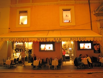 A warmly lit outdoor cafe scene with a gelateria, wine bar, and bistrot. Patrons sit at small tables under a large awning, while the interior glows with inviting lights. The facade is painted in shades of orange and beige, and there are two windows on the upper floor.