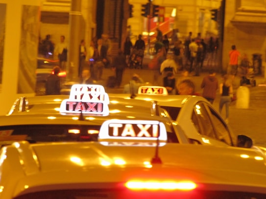 Several taxi cars are aligned in a busy urban street at night, with illuminated taxi signs on their roofs. People are walking in the background, suggesting a bustling nightlife atmosphere. The ambient lighting is warm, casting a yellowish glow over the scene.