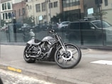 A sleek black motorcycle is parked on a sidewalk in an urban area. The background features a modern building with large reflective windows and a few cars parked on the street. The motorcycle has a low-slung design with a prominent engine and streamlined appearance.