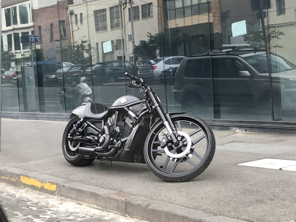 A sleek black motorcycle is parked on a sidewalk in an urban area. The background features a modern building with large reflective windows and a few cars parked on the street. The motorcycle has a low-slung design with a prominent engine and streamlined appearance.