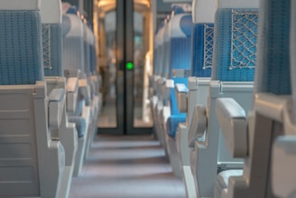A view down the aisle of a train carriage with rows of empty seats upholstered in blue fabric. The seats have armrests and are arranged in a symmetrical layout. Soft lighting creates a calm atmosphere, and the back of the carriage is visible with a door leading to another section.