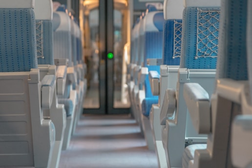 A view down the aisle of a train carriage with rows of empty seats upholstered in blue fabric. The seats have armrests and are arranged in a symmetrical layout. Soft lighting creates a calm atmosphere, and the back of the carriage is visible with a door leading to another section.