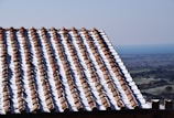 A sunlit roof covered with rows of terracotta tiles, partially dusted with snow, with a distant view of a coastal landscape under a clear blue sky.