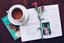 A cup of tea on a wooden table, next to an open magazine with text and an image of a person in pink. A small bouquet of pink flowers lies on top of the magazine, and a green book titled 'seeds planted in concrete' rests partially underneath.
