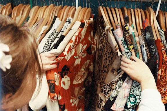 A person is browsing through a variety of colorful and patterned clothes on hangers. The clothing includes floral prints, leopard patterns, and other vibrant designs. The hangers are wooden, and the person's hands are actively sorting through the selection.
