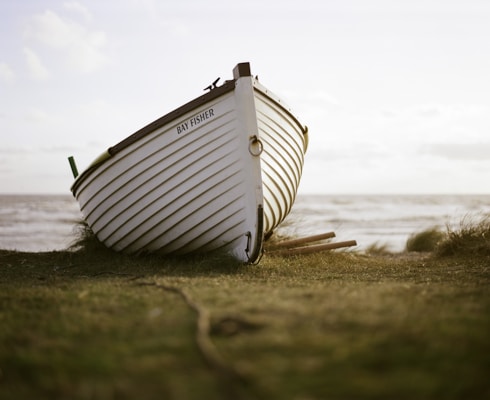 A white wooden boat rests on a grassy shore with the ocean in the background. The boat is titled 'Bay Fisher' and its worn surface suggests frequent use. The horizon shows a slightly cloudy sky, and the lighting indicates late afternoon or early morning.