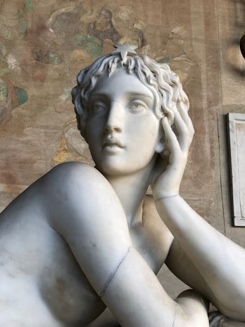 A marble sculpture of a person with an intricate hairstyle, resting their head on one hand while the other arm wraps around the chest. The background contains faded frescoes with earthy tones.