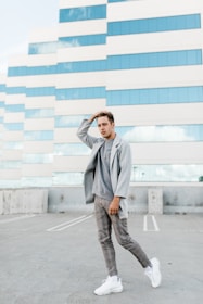 A person dressed in a casual grey blazer and matching trousers stands confidently on a rooftop parking lot. The backdrop features a modern building with a grid-like pattern of windows and beige panels reflecting the sky.