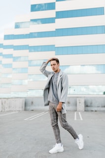A person dressed in a casual grey blazer and matching trousers stands confidently on a rooftop parking lot. The backdrop features a modern building with a grid-like pattern of windows and beige panels reflecting the sky.
