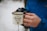 A person wearing a blue jacket is holding a white enamel mug with black lettering that reads 'The Adventure Begins.' The person is also holding a camera, which is partially visible in the background. The setting appears to be outdoors, with snow visible in the background.