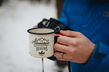 A person wearing a blue jacket is holding a white enamel mug with black lettering that reads 'The Adventure Begins.' The person is also holding a camera, which is partially visible in the background. The setting appears to be outdoors, with snow visible in the background.
