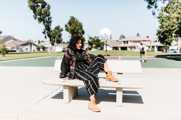 A person is sitting casually on a bench in an outdoor area with a basketball court in the background. They are dressed in a stylish outfit featuring a black jacket, black striped pants, and tan boots. The setting is bright and sunny, with clear skies and residential houses visible in the distance. Another person is seen in the background holding a basketball.