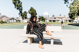 A person is sitting casually on a bench in an outdoor area with a basketball court in the background. They are dressed in a stylish outfit featuring a black jacket, black striped pants, and tan boots. The setting is bright and sunny, with clear skies and residential houses visible in the distance. Another person is seen in the background holding a basketball.