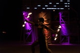 A performer is skillfully manipulating fire in a dimly lit environment, creating a dynamic and captivating display. In the background, vibrant purple and orange lights illuminate the scene, enhancing the dramatic atmosphere.