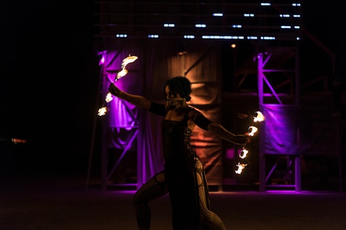A performer is skillfully manipulating fire in a dimly lit environment, creating a dynamic and captivating display. In the background, vibrant purple and orange lights illuminate the scene, enhancing the dramatic atmosphere.