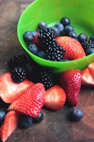 A variety of fresh berries, including strawberries, blueberries, and blackberries, are placed in and around a bright green bowl. The berries appear vibrant and ripe, with a glossy sheen on the blackberries indicating freshness. The background is a textured brown surface that highlights the colors of the berries.
