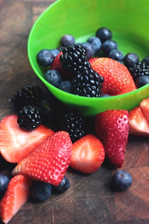 A variety of fresh berries, including strawberries, blueberries, and blackberries, are placed in and around a bright green bowl. The berries appear vibrant and ripe, with a glossy sheen on the blackberries indicating freshness. The background is a textured brown surface that highlights the colors of the berries.