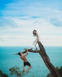 woman sitting on tree trunk with man holding on branch near sea under white clouds during daytime
