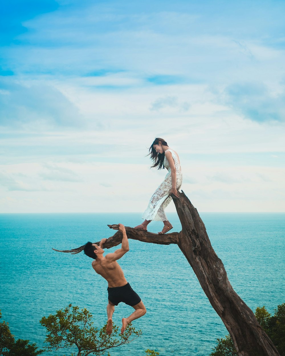 woman sitting on tree trunk with man holding on branch near sea under white clouds during daytime