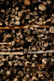 A tightly packed stack of chopped firewood, featuring various sizes and shapes of logs. The wood displays natural textures and tones, with cut surfaces and bark visible.