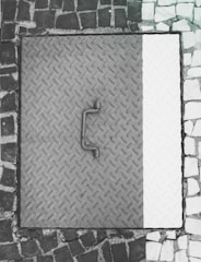 A metal manhole cover with a textured diamond plate pattern is surrounded by a mosaic of dark and light cobblestones. The cover appears worn with a handle in the center.