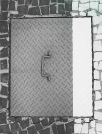 A metal manhole cover with a textured diamond plate pattern is surrounded by a mosaic of dark and light cobblestones. The cover appears worn with a handle in the center.