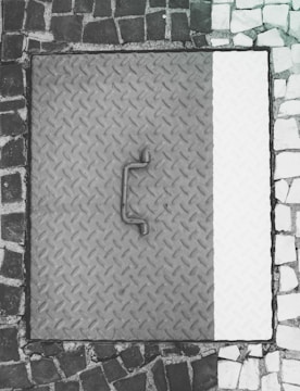 A metal manhole cover with a textured diamond plate pattern is surrounded by a mosaic of dark and light cobblestones. The cover appears worn with a handle in the center.
