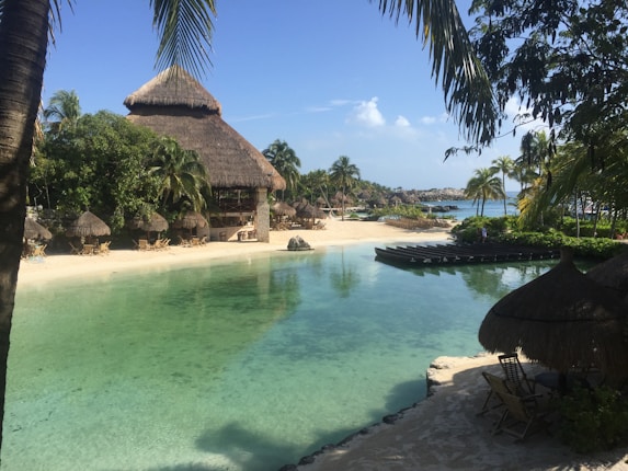 A tranquil tropical scene featuring a clear lagoon bordered by sandy beaches and lush greenery. Thatched-roof huts are scattered along the shoreline, and palm trees sway gently in the breeze. The water is a vibrant turquoise, and the sky is mostly clear with a few clouds.