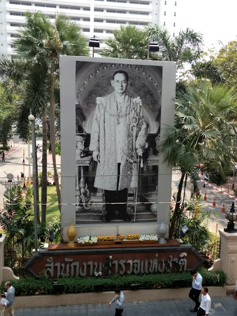 A large portrait of a man in formal attire is prominently displayed in an outdoor setting. The portrait is surrounded by a decorative frame, and there are several lit lamps in the vicinity. Behind the portrait are a variety of tropical plants and a white multi-story building. Below the portrait, there is a decorative arrangement with flowers and ceremonial objects. Pedestrians are walking by on the sidewalk in front of the scene.