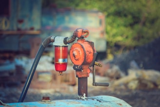 An old, rusted industrial valve is attached to a pipe. A red cylindrical component, possibly a filter or gauge, is connected to the valve with a flexible hose. The background appears blurred, featuring greenish hues indicating foliage or outdoor surroundings.