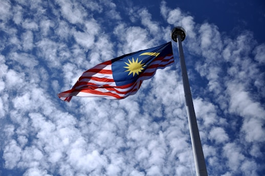 A Malaysian flag with vibrant red and white stripes and a blue canton with a yellow crescent and star flows majestically against a bright, cloud-filled sky. The flagpole extends high into the air, making the flag prominent against the scattered clouds.