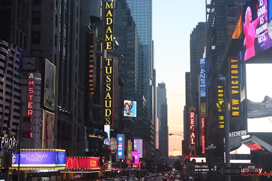 A bustling city street lined with tall buildings, each adorned with vibrant neon signs advertising theaters and attractions. The sun is setting in the background, casting a warm glow over the scene. Crowds and traffic fill the street, adding to the dynamic urban atmosphere.