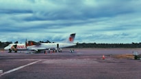 An airplane is parked on an airport runway with ground crew members attending to it. The sky is overcast, and there are scattered clouds. Orange traffic cones are set around the aircraft, and a small baggage cart is visible nearby. The surrounding area is lined with trees in the distance.