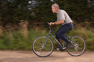 An elderly man rides a bicycle along a path with a blurred background, suggesting motion. He wears a striped shirt and dark pants, with glasses and a wristwatch.