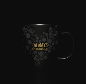 A dark-colored mug with floral and circular decorative patterns, featuring the words 'Always dreaming' in yellow text.