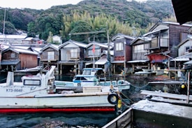 A picturesque village scene with traditional Japanese wooden houses built on stilts along a calm waterfront. Several small boats are docked close to the houses, and lush green hills rise in the background. The area appears serene and lightly dusted with snow, suggesting a cold season.