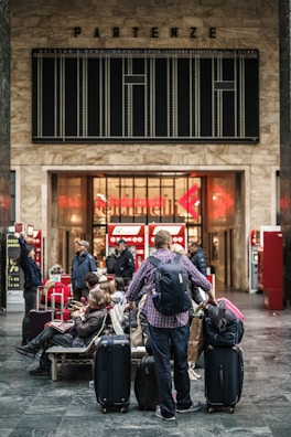 A busy train station with several travelers carrying luggage. People are sitting and standing in front of ticket machines. The station features a large departure board above the entrance with an old-style design and a 'Partenze' sign, indicating departures.