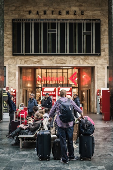 A busy train station with several travelers carrying luggage. People are sitting and standing in front of ticket machines. The station features a large departure board above the entrance with an old-style design and a 'Partenze' sign, indicating departures.