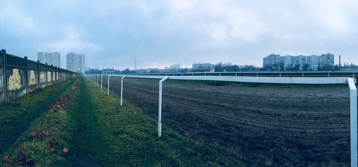 A wide view of a racetrack with a dirt path to the right and a grassy path to the left. A long fence runs parallel to the track on the left. The background features several large residential buildings under a cloudy sky, creating an urban landscape.