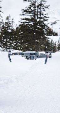 A drone hovers in a snowy landscape with tall evergreen trees in the background. The scene is characterized by the white snow covering the ground and the overcast sky, creating a serene winter atmosphere.