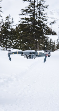 A drone hovers in a snowy landscape with tall evergreen trees in the background. The scene is characterized by the white snow covering the ground and the overcast sky, creating a serene winter atmosphere.