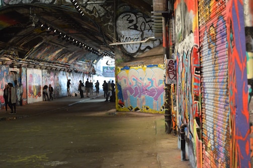 A graffiti-covered urban tunnel with colorful artwork on the walls. People are gathered and walking through the tunnel, casting shadows on the ground. The walls and ceiling are densely decorated with vibrant street art and graffiti.