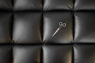 A pair of scissors lies on a black, tufted leather surface. The stitching creates a quilted pattern, adding texture to the material and reflecting a slight sheen.