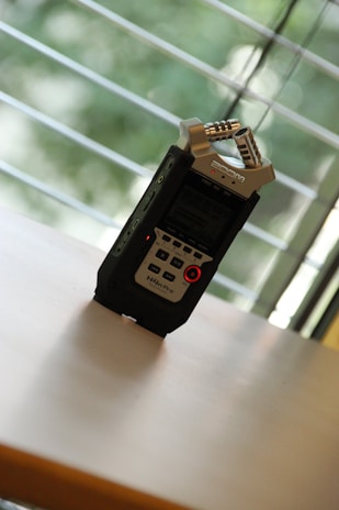 A portable audio recorder is placed on a smooth flat surface, with a background consisting of a window with horizontal blinds. The recorder is black with metallic elements, featuring multiple buttons and a digital display. The recorder is angled slightly, capturing natural light filtering through the blinds.