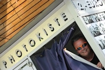 A man with sunglasses peeks out from behind the curtain of a photo booth. The booth has a sign labeled 'PHOTOKISTE' and another indicating a cost of 2 euros. There's a display of black and white photos pinned on the side of the booth.