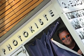 A man with sunglasses peeks out from behind the curtain of a photo booth. The booth has a sign labeled 'PHOTOKISTE' and another indicating a cost of 2 euros. There's a display of black and white photos pinned on the side of the booth.