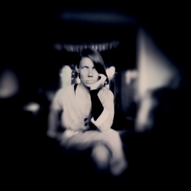 A monochrome image with high contrast depicting a person sitting with a hand supporting their chin in a contemplative pose. The foreground is sharply focused while the background is blurred, creating a sense of depth.
